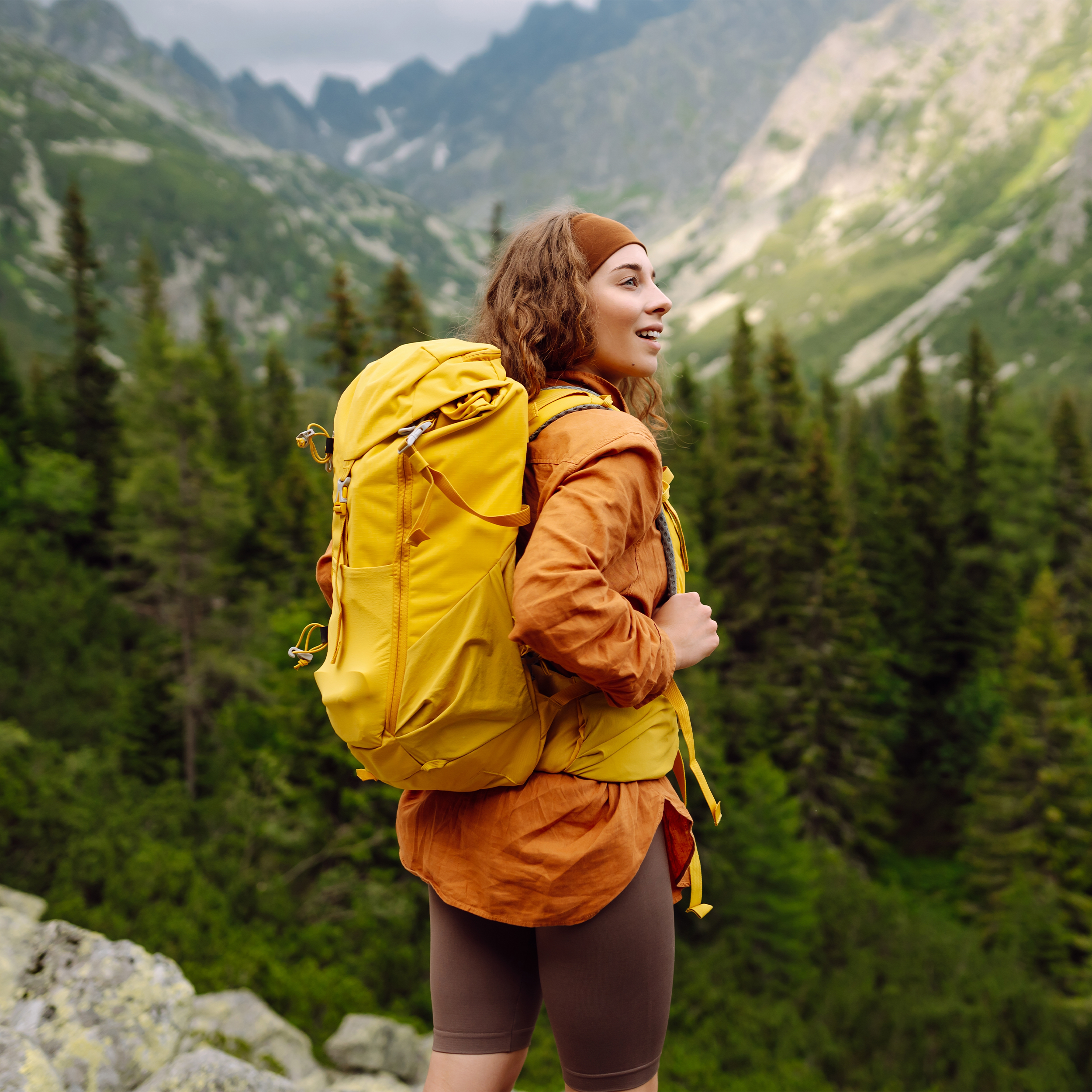 Eine junge Frau steht mit Wanderkleidung und einem gelben Rucksack in der Natur und bewundert die Landschaft.