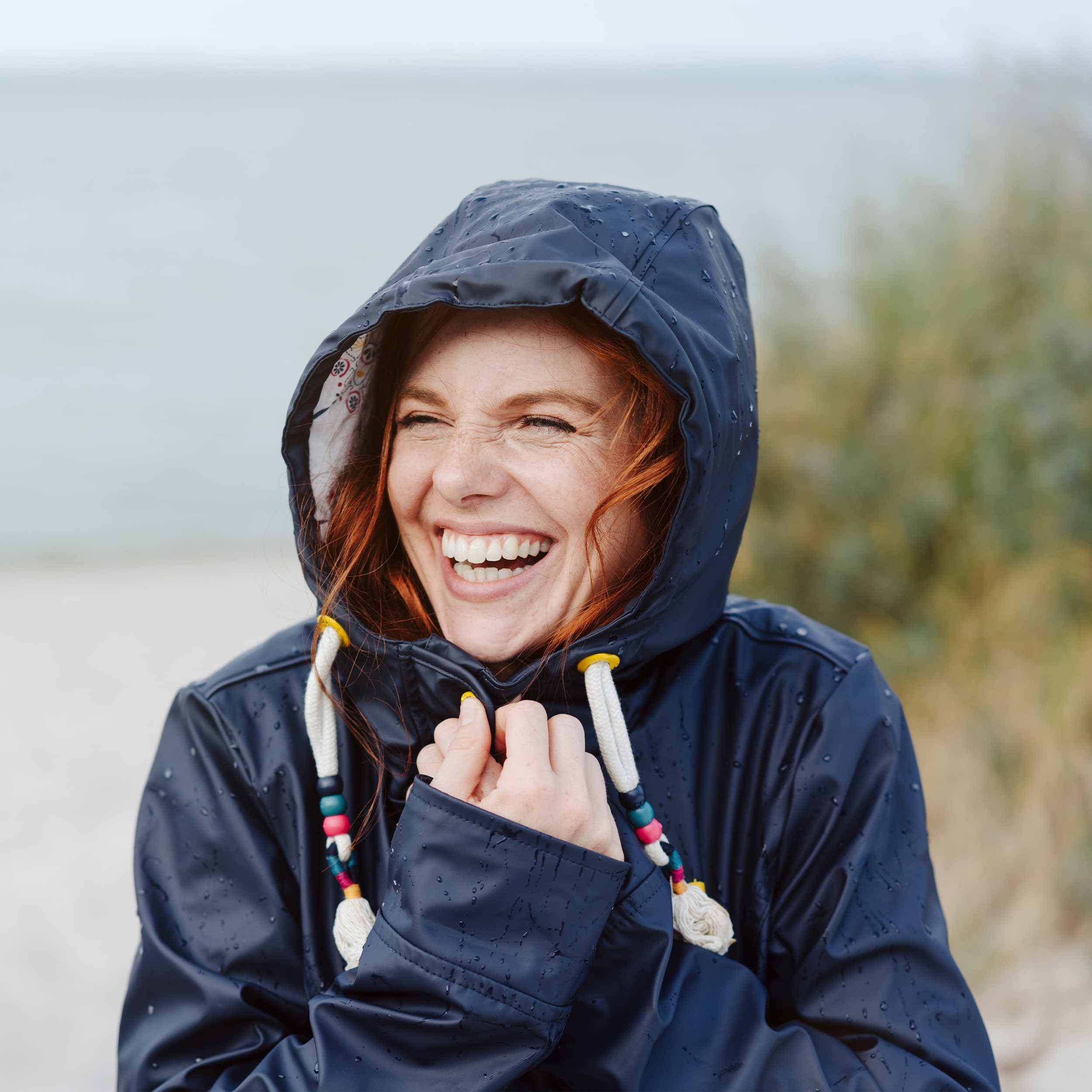 Eine rothaarige Frau in den Dreissigern sitzt in der Natur an einem Strand. Sie trägt eine Regenjacke inklusive Kapuze und hält diese vorne fest. Es ist windig. Die Frau lacht.