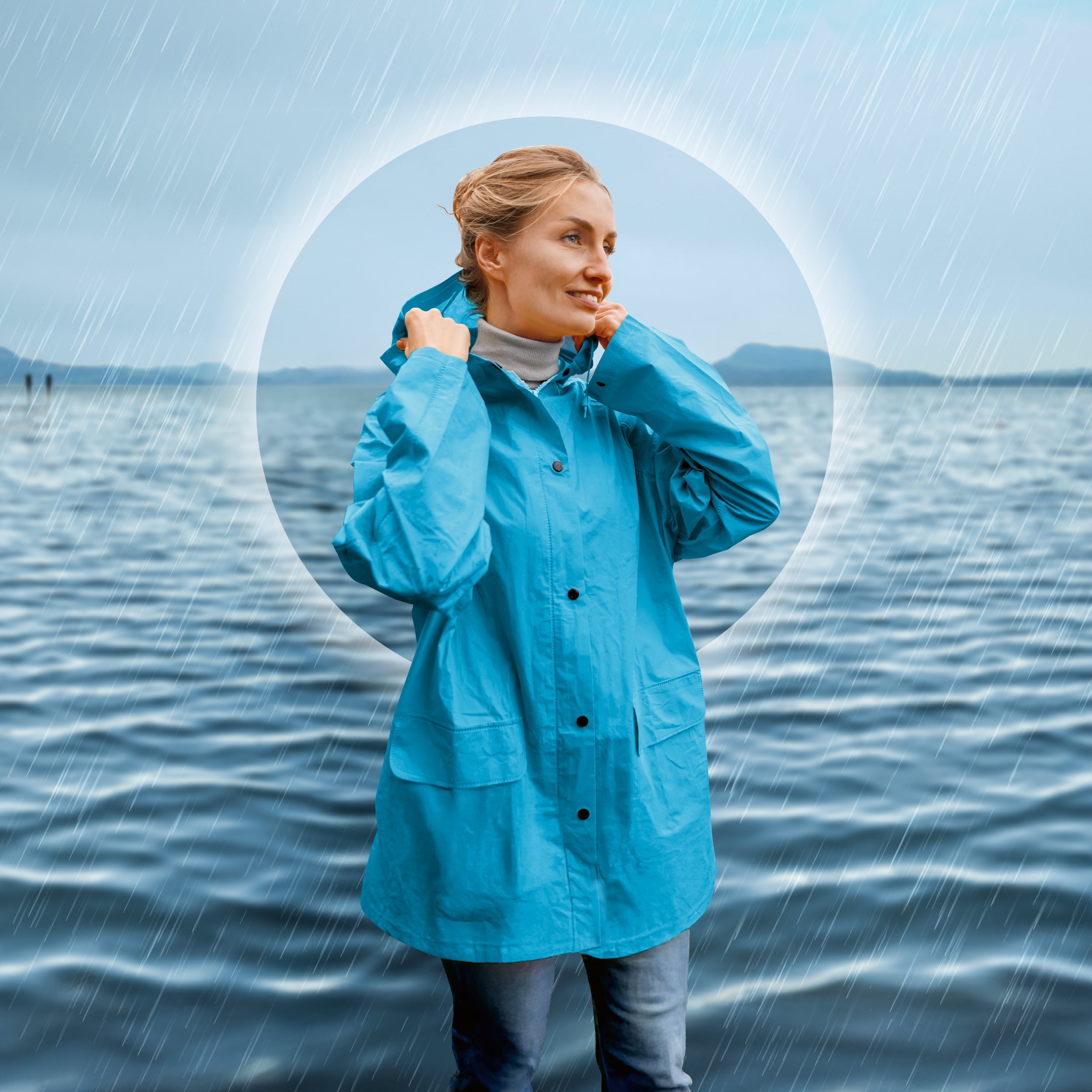 Eine Frau steht mit einer blauen Regenjacke bei einem Gewässer. Sie hält die Kapuze mit den Händen fest, schaut in die Ferne und lächelt.