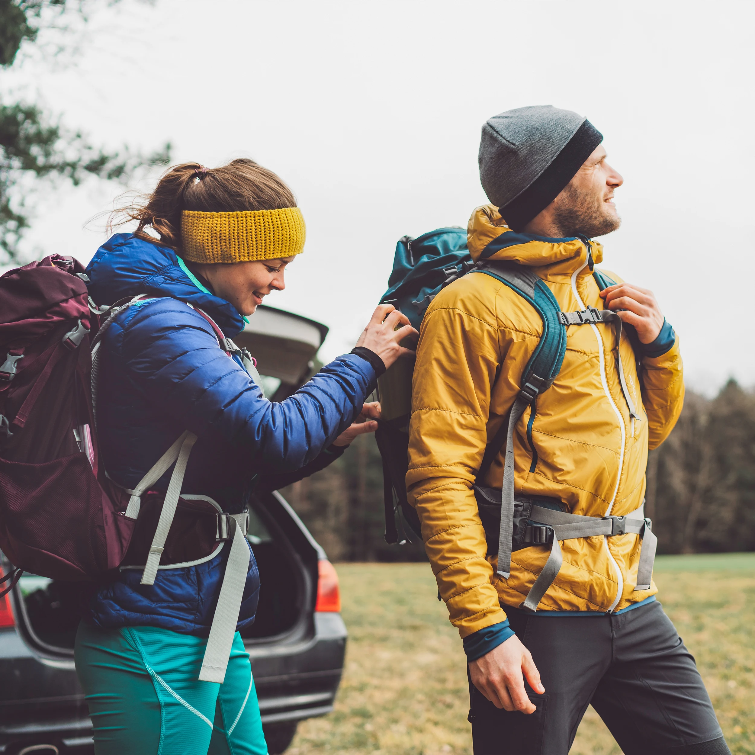 Ein junge Frau und ein junger Mann, wahrscheinlich ein Paar, stehen in Wanderausrüstung vor einem Auto mit offenem Kofferraum. Die Frau verstellt etwas am Rucksack des Mannes. Beide sehen glücklich aus.